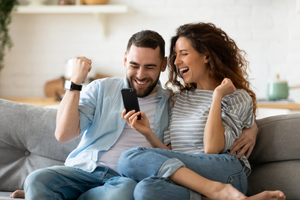 Excited,Overjoyed,Couple,Resting,On,Couch,Holding,Smart,Phone,Celebrating